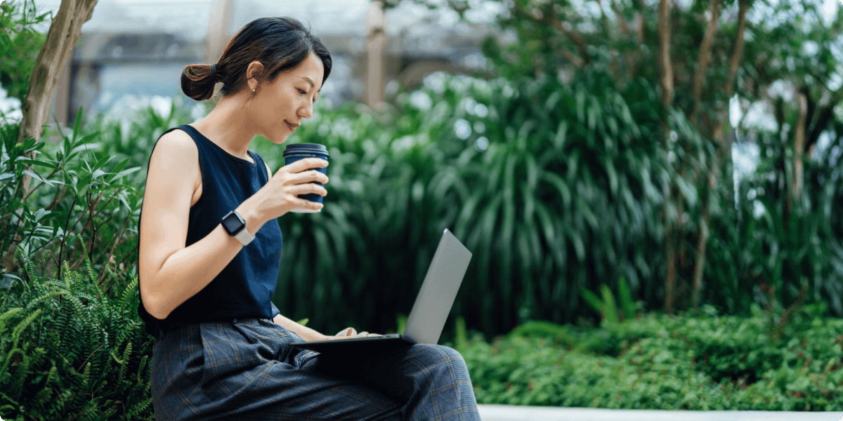 Eine Frau arbeitet draußen auf ihrem Laptop und hält dabei einen Becher mit Kaffee. Sie ist von saftigem Grün umgeben.