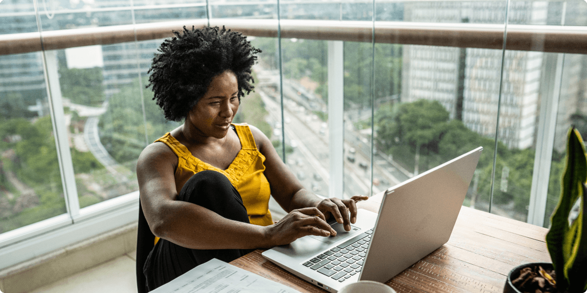 Uma mulher trabalhando em seu laptop na varanda de seu apartamento. Há uma vista de árvores e apartamentos altos atrás dela.